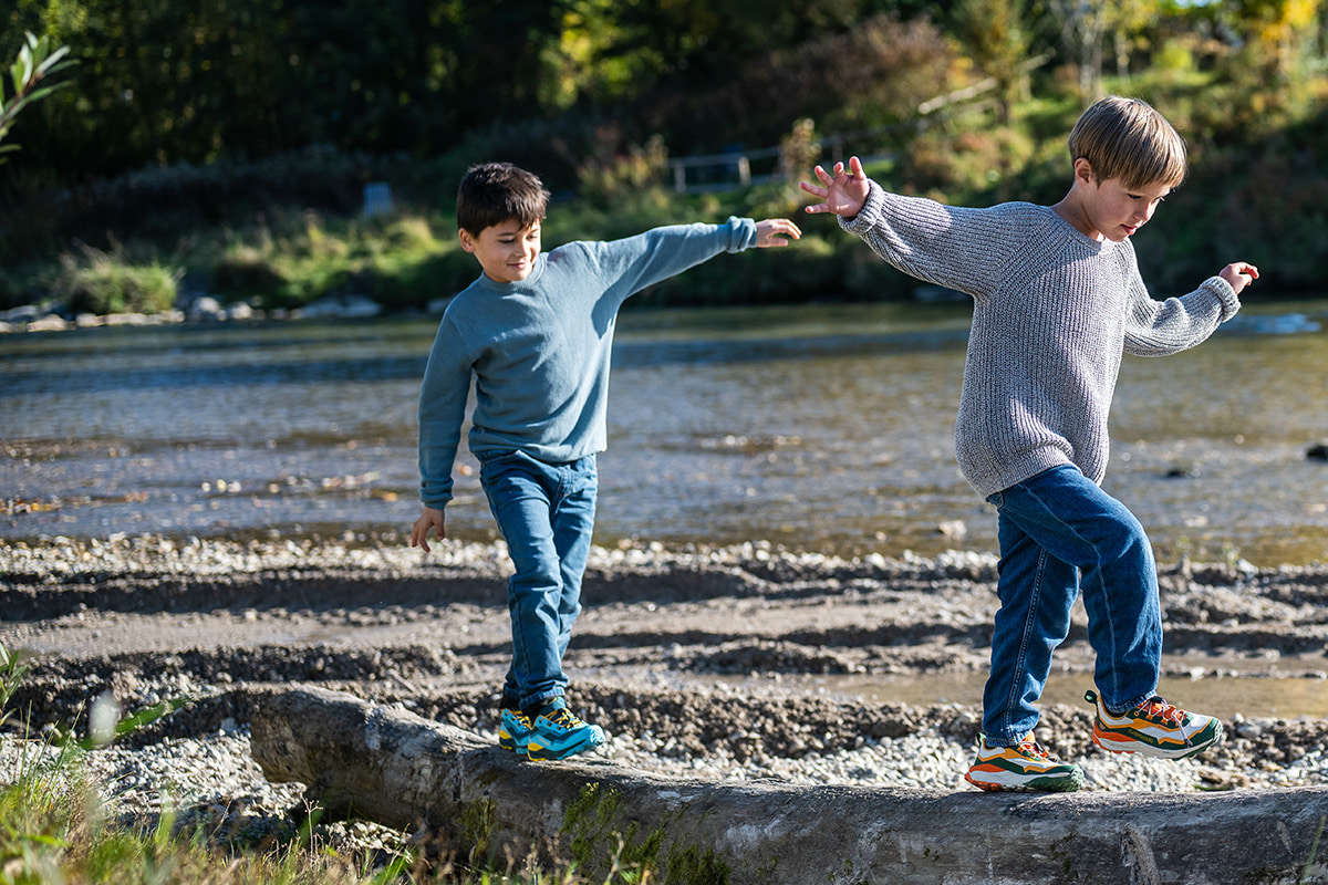 Two boys balance on a tree trunk by the riverbank, playing happily outdoors.