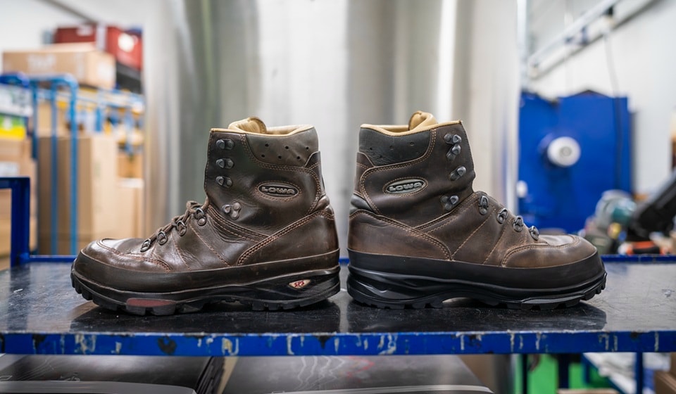 Two used brown Lowa hiking boots stand on a blue shelf in a workshop. The shoes are made of leather and show signs of wear. The background is blurred and shows workshop equipment.